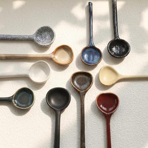 以下が含まれることがあります： 青、ベージュ、黒、茶色など、さまざまな色のセラミックスプーンのコレクション。 スプーンは長い柄と丸みを帯びたボウルがあり、スープやソースの提供に最適です。 スプーンは白い表面に配置されています。