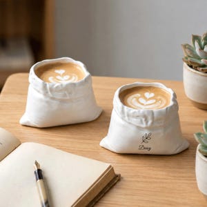 May include: Two white ceramic mugs shaped like small, folded fabric sacks, each filled with coffee featuring heart-shaped latte art. The mug on the right features a small black line drawing of a sprig and the name Daisy printed in black text.