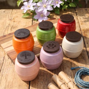 May include: A collection of ceramic pet urns in various colors, including orange, green, red, white, and pink. Each urn has a dark brown lid with an engraved design and the name "Coco" or the phrase "We miss you".
