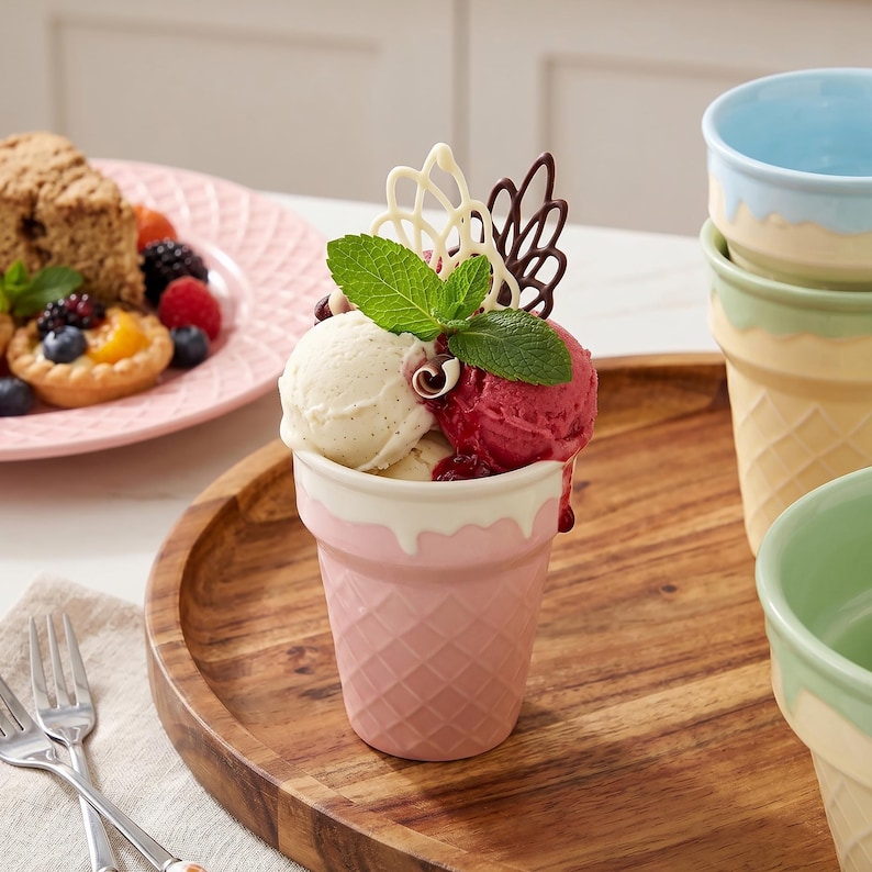 May include: A pink ceramic ice cream cup filled with vanilla and raspberry ice cream, topped with mint leaves and chocolate decorations. The cup has a waffle cone design and is part of a set with other pastel-colored cups.
