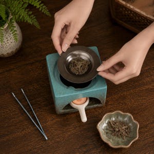 May include: A square teal ceramic incense burner with a dark brown metal tray containing dried herbs, a small white ceramic candle holder with a lit tea light, a pair of thin metal tweezers, and a small scalloped edge ceramic dish with dried herbs.
