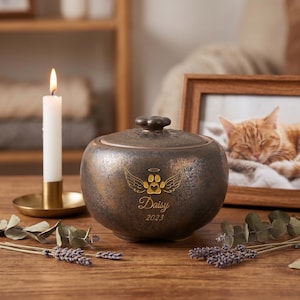 Könnte beinhalten: Bronze-Haustierurne mit Deckel, Pfotenabdruck-Design mit Engelsflügeln, dem Namen "Daisy" und dem Jahr 2023. Eine brennende Kerze in einem goldenen Halter und ein gerahmtes Foto einer Katze im Hintergrund.