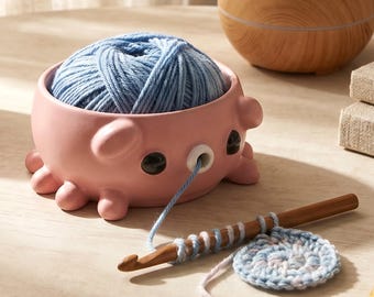 May include: A pink ceramic yarn bowl shaped like an octopus with black eyes and a small hole for yarn, containing a ball of light blue yarn. A wooden crochet hook with a partially worked light blue circular crochet piece is positioned in front.