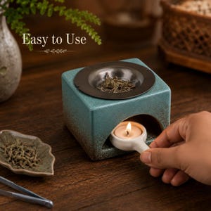 May include: A square teal ceramic oil burner with a dark brown gradient base and a metal top dish containing dried herbs. The text "Easy to Use" is visible above. A small white ceramic handle holds a lit tea light candle inside the burner.