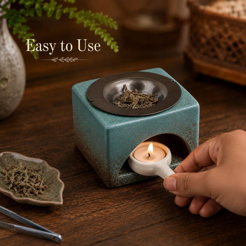 May include: A square teal ceramic oil burner with a dark brown gradient base and a metal top dish containing dried herbs. The text "Easy to Use" is visible above. A small white ceramic handle holds a lit tea light candle inside the burner.