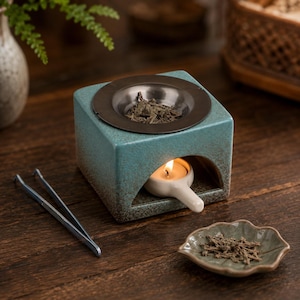 May include: A square teal ceramic incense burner with a dark brown gradient base and a circular metal tray on top containing dried herbs. A small white tea light candle sits in the arched opening, and metal tweezers rest nearby.