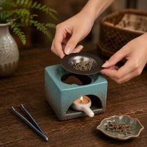 May include: A square teal ceramic incense burner with a speckled brown base, featuring a central circular opening for a metal tray. A small white ceramic candle holder with a lit tea light sits inside the burner, accompanied by metal tweezers.