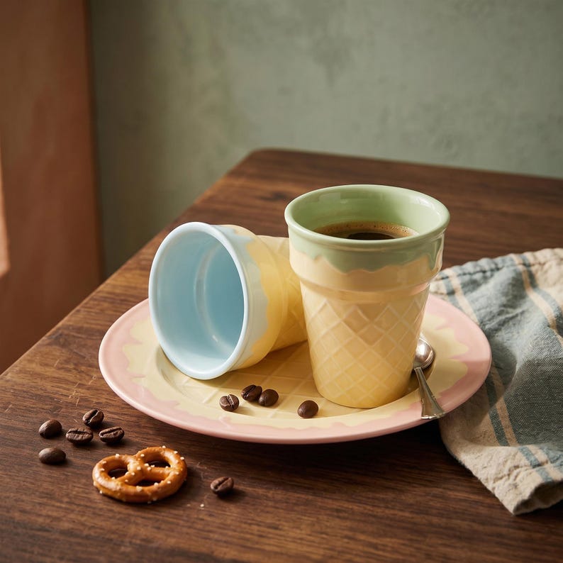 May include: Two ice cream cone-shaped coffee mugs, one filled with coffee, sit on a plate with a pink and yellow rim. A pretzel and coffee beans are on the wooden table. The mugs have a waffle cone texture.