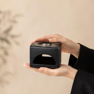 May include: A matte black ceramic oil burner featuring a cubic shape with a wide, arched opening at the base for a tea light candle and a circular, recessed basin at the top for holding essential oils or wax melts in a solid, dark finish.