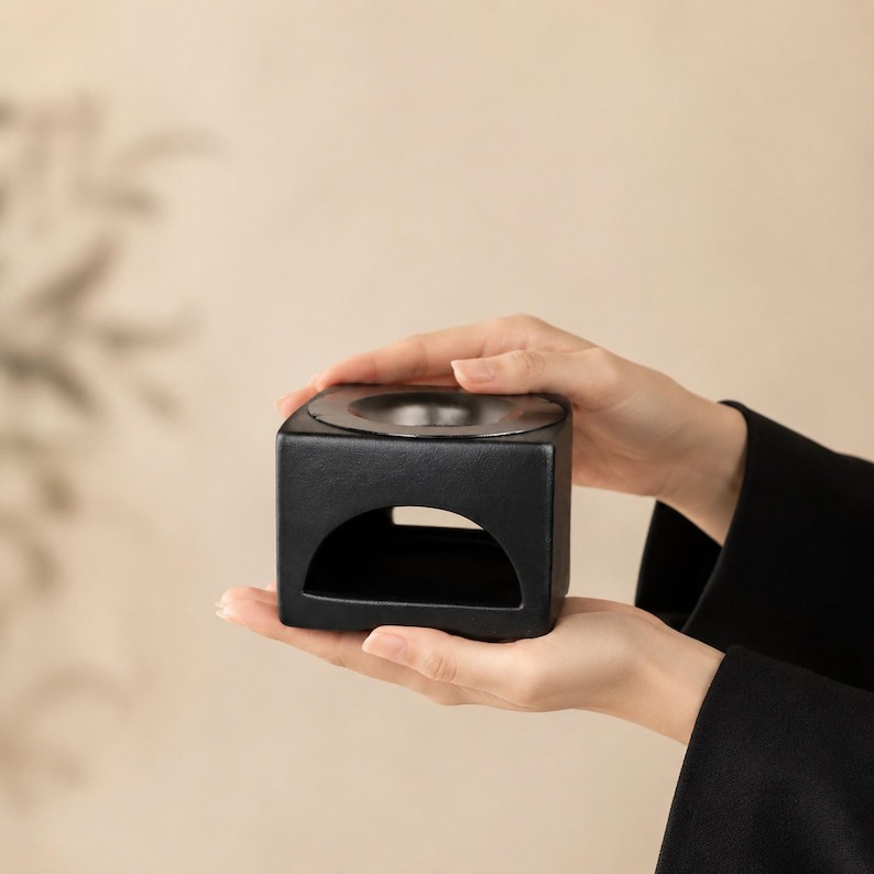 May include: A matte black ceramic oil burner featuring a cubic shape with a wide, arched opening at the base for a tea light candle and a circular, recessed basin at the top for holding essential oils or wax melts in a solid, dark finish.