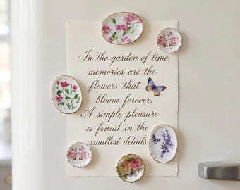 May include: A collection of small, decorative floral plates with gold rims, arranged around a piece of paper with calligraphy. The paper has a butterfly illustration and the text: "In the garden of time, memories are the flowers that bloom forever."