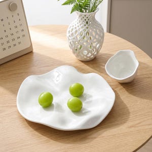 May include: A white, organically shaped ceramic plate holds three green grapes. A small white bowl and a decorative white vase with a lattice design are also present. A calendar is visible in the background. The items are on a light wood surface.