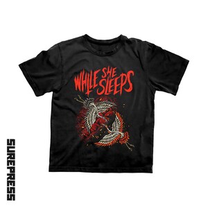 May include: A black short-sleeve t-shirt featuring the red text While She Sleeps above a graphic of two cranes flying in a circular motion. The cranes are white and red with detailed feathers, set against a dark red circular background with small sparks.
