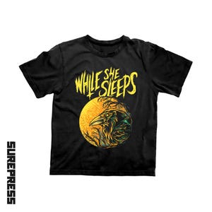 May include: A black short-sleeve t-shirt featuring the yellow text WHILE SHE SLEEPS above a graphic of a raven perched in front of a large yellow moon with dark tree branches. The word SUREPRESS is printed vertically in black on the left side.