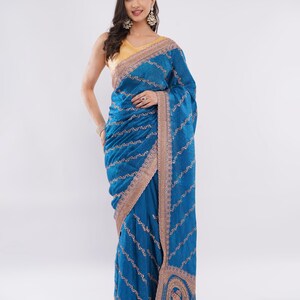 Peut inclure: Une femme porte un sari bleu vif avec des broderies dorées complexes. Le sari présente une bordure détaillée et un motif décoratif à l'ourlet. Le chemisier est de couleur jaune clair.
