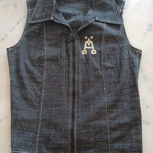 Könnte beinhalten: Eine ärmellose Jeansweste in Dunkelgrau mit Kragen und durchgehendem schwarzem Reißverschluss. Weiße Nähte umranden die Nähte und den Kragen. Ein weißes Emblem befindet sich auf der Vorderseite. Die Weste hat eine taillierte Passform.