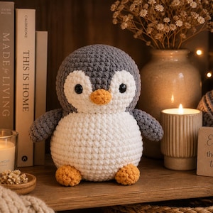 Op de afbeelding: Een handgemaakte gehaakte pinguïn met een grijze kop en vleugels, een witte buik en een oranje snavel en voeten. De pinguïn staat op een houten plank naast een brandende kaars, boeken en een vaas met bloemen. Een klein houten bordje leest "Enjoy the little things."
