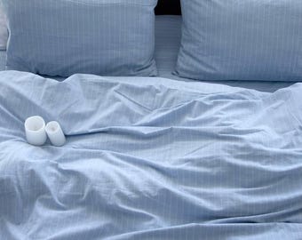 Może przedstawiać: Łóżko z jasnoniebieską pościelą i dwiema pasującymi poszewkami na poduszki. Pościel ma subtelny wzór w paski. Na łóżku znajdują się dwie małe, białe, cylindryczne świece. Tło jest ciemne.