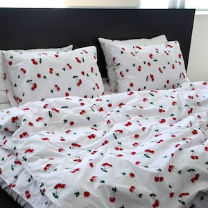 May include: White bedding set with a repeating pattern of red cherries and green stems. The set includes two pillowcases and a duvet cover, all with the same cherry design. The bed has a black headboard.