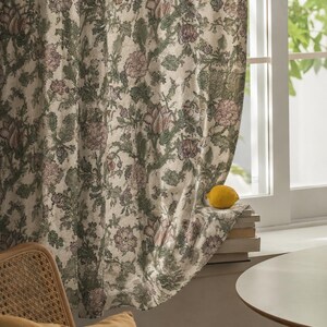 May include: A floor-length curtain featuring a dense botanical print with muted green foliage and soft pink floral blossoms. The fabric hangs in vertical folds, partially covering a window frame where a single yellow lemon rests on a stack of books.