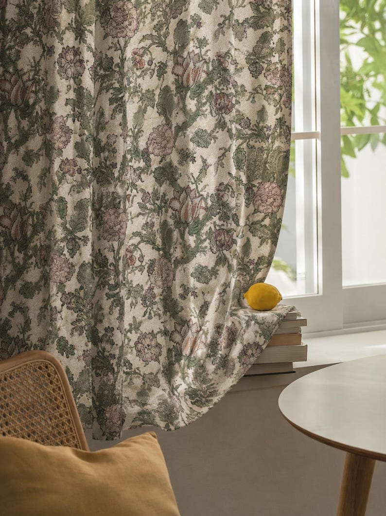 May include: A floor-length curtain featuring a dense botanical print with muted green foliage and soft pink floral blossoms. The fabric hangs in vertical folds, partially covering a window frame where a single yellow lemon rests on a stack of books.