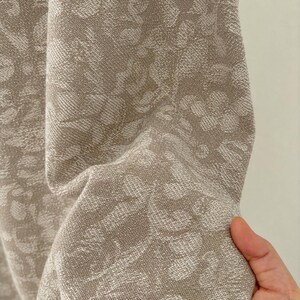 Può includere: Un tessuto beige e bianco caratterizzato da un fitto motivo floreale intrecciato. Il materiale presenta una superficie strutturata con un design botanico ripetitivo in tonalità neutre chiare, che mette in risalto l'intreccio complesso e l'interazione tra i due colori.