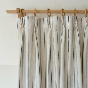 K&ouml;nnte beinhalten: Ein beigefarbener Vorhang mit vertikalen marineblauen Streifen und Faltenwurf am oberen Rand. Der Vorhang ist an einer hellen Holzstange mit passenden Holzringen befestigt, was f&uuml;r einen strukturierten und gleichm&auml;&szlig;igen Fall des Stoffes sorgt.