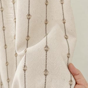 Puede incluir: Una tela de color crema con rayas verticales compuestas por pequeños detalles copetudos en forma de diamante en un tono marrón claro. El tejido tiene una textura visible y está drapeado para mostrar su calidad y flexibilidad.