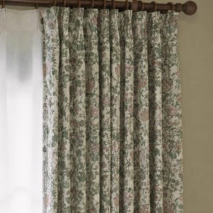 May include: Pleated curtains featuring a floral pattern in muted shades of green, pink, and cream, suspended from a dark brown wooden rod with rings. A sheer white curtain panel is visible on the left side, hanging from the same wooden rod structure.