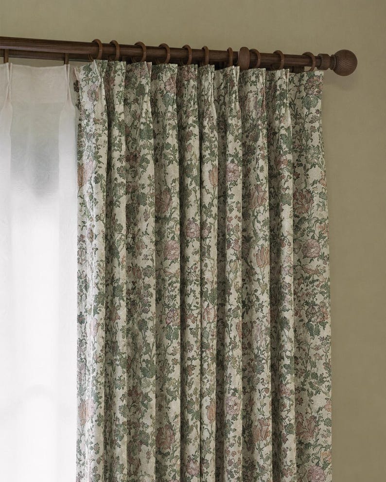 May include: Pleated curtains featuring a floral pattern in muted shades of green, pink, and cream, suspended from a dark brown wooden rod with rings. A sheer white curtain panel is visible on the left side, hanging from the same wooden rod structure.