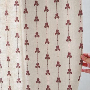 May include: A beige fabric curtain featuring a repeating vertical pattern of dark red floral motifs connected by thin lines. The textile has a visible woven texture, and the design consists of small, stylized blossoms arranged in straight columns.