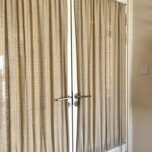 May include: Two beige curtains with a thin yellow grid pattern hanging over a set of white double doors. The curtains are gathered at the top and bottom, featuring a textured fabric finish and silver-toned metal door handles visible in the centre.