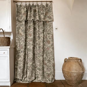 May include: A floor-length curtain featuring a dense floral pattern in muted shades of green, brown, and cream. The fabric is suspended from a thin metal rod by small circular rings and includes an integrated valance with a scalloped bottom edge.