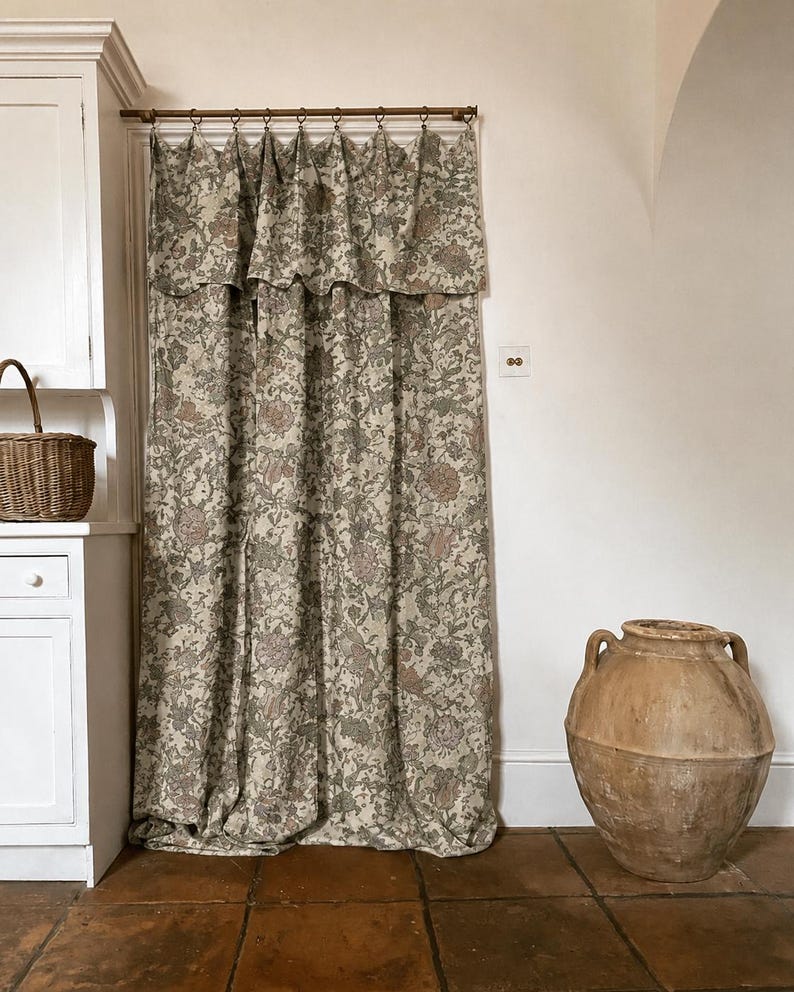 May include: A floor-length curtain featuring a dense floral pattern in muted shades of green, brown, and cream. The fabric is suspended from a thin metal rod by small circular rings and includes an integrated valance with a scalloped bottom edge.
