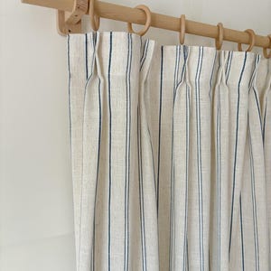 K&ouml;nnte beinhalten: Cremefarbene Vorh&auml;nge mit vertikalen marineblauen Nadelstreifen, aufgeh&auml;ngt an einer hellen Holzstange mit passenden Holzringen. Der Stoff weist ein strukturiertes Gewebe auf und ist an der Oberkante des Vorhangs in Falten gelegt.