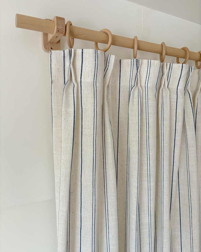 K&ouml;nnte beinhalten: Cremefarbene Vorh&auml;nge mit vertikalen marineblauen Nadelstreifen, aufgeh&auml;ngt an einer hellen Holzstange mit passenden Holzringen. Der Stoff weist ein strukturiertes Gewebe auf und ist an der Oberkante des Vorhangs in Falten gelegt.