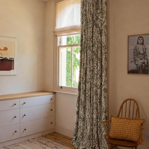 May include: Floor-to-ceiling window curtains featuring a dense botanical print in muted green and brown tones on a cream background, paired with a sheer white Roman shade covering the upper portion of the window frame.
