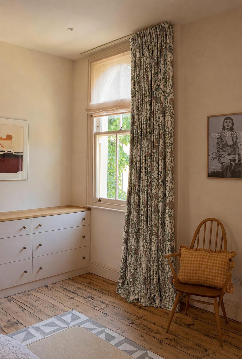 May include: Floor-to-ceiling window curtains featuring a dense botanical print in muted green and brown tones on a cream background, paired with a sheer white Roman shade covering the upper portion of the window frame.