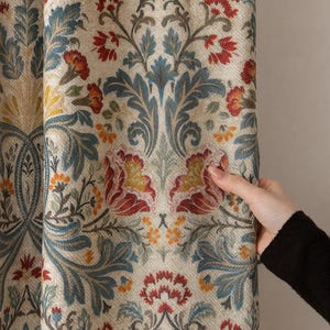 Op de afbeelding: Close-up van een gordijn met bloemenpatroon. Het gordijn heeft een herhalend ontwerp van rode, blauwe en gele bloemen en bladeren op een beige achtergrond. De stof heeft een getextureerde uitstraling.