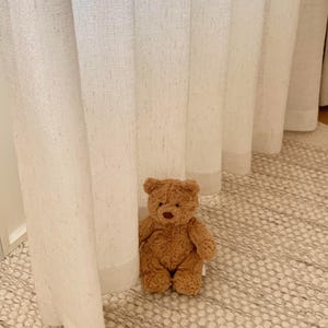 May include: A small, plush teddy bear with curly, light brown fur, a dark brown stitched nose, and rounded ears. The bear is seated in an upright position, featuring a soft, textured appearance with visible seams and a simple, classic design.