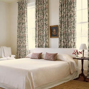 May include: A bedroom featuring a bed with a cream-colored coverlet and two rectangular pillows with a red and cream pattern. Long curtains with a green and pink floral print hang over windows. A small wooden table holds a lamp and flowers.