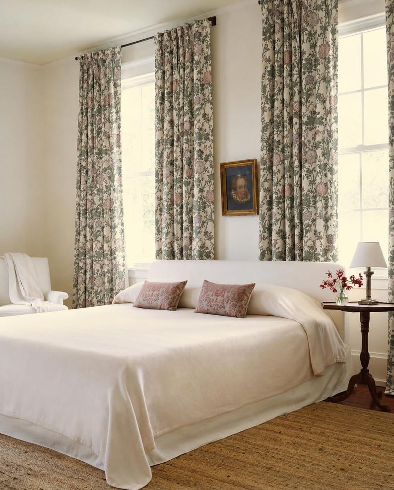 May include: A bedroom featuring a bed with a cream-colored coverlet and two rectangular pillows with a red and cream pattern. Long curtains with a green and pink floral print hang over windows. A small wooden table holds a lamp and flowers.