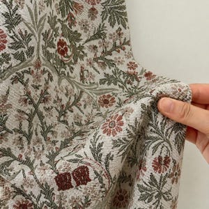 Könnte beinhalten: Ein gewebter Stoff mit einem dichten botanischen Muster aus komplexen Blumen- und Blattmotiven in gedeckten Rot-, Salbeigrün- und Cremetönen. Das Textil weist eine strukturierte Oberfläche mit einem sich wiederholenden Design aus Blumen und Laub auf.