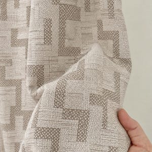 Può includere: Un tessuto intrecciato caratterizzato da un motivo geometrico di blocchi a forma di L incastrati tra loro in tonalità beige e crema. Il tessuto presenta una trama visibile con una struttura a griglia coerente che crea un motivo ripetitivo.