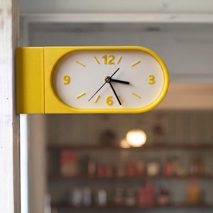Puede incluir: Un reloj ovalado montado en la pared con un marco amarillo brillante y una esfera blanca. El reloj presenta números amarillos en relieve en las posiciones de las 3, 6, 9 y 12, con marcas amarillas para las horas restantes y manecillas negras.