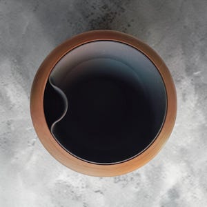 Pode incluir: Vista aérea de um recipiente de metal cor de cobre com interior escuro. O recipiente tem formato arredondado com superfície lisa e reflexiva. O interior é preto e uma reentrância curva é visível na lateral.