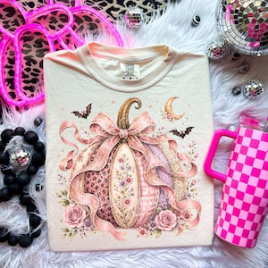 May include: A cream-colored t-shirt featuring a central graphic of a pumpkin decorated with pink ribbons, floral patterns, leopard print, and plaid sections. The design includes small bats, a crescent moon, and pink roses at the base of the pumpkin.