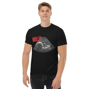 Könnte beinhalten: Schwarzes T-Shirt mit einem Graustufen-Ultraschallbild und dem Text "BORN TO READ" in einem roten Feld. Das Bild zeigt einen Fötus und medizinische Bildgebung. Das T-Shirt hat einen klassischen Rundhalsausschnitt.