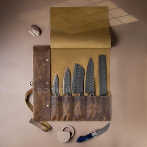 Pode incluir: Um estojo de couro castanho para facas contendo cinco facas de cozinha com lâminas de metal escuro martelado. O estojo apresenta o texto BORITH STUDIO. Uma faca separada com cabo azul e lâmina curva encontra-se abaixo, junto a um objeto circular.