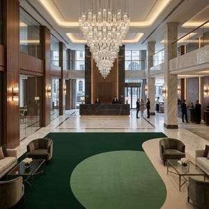 May include: A grand hotel lobby featuring a large, ornate chandelier suspended above a dark marble reception desk. The space includes a green and beige patterned carpet, seating areas, and large windows, creating a luxurious atmosphere.